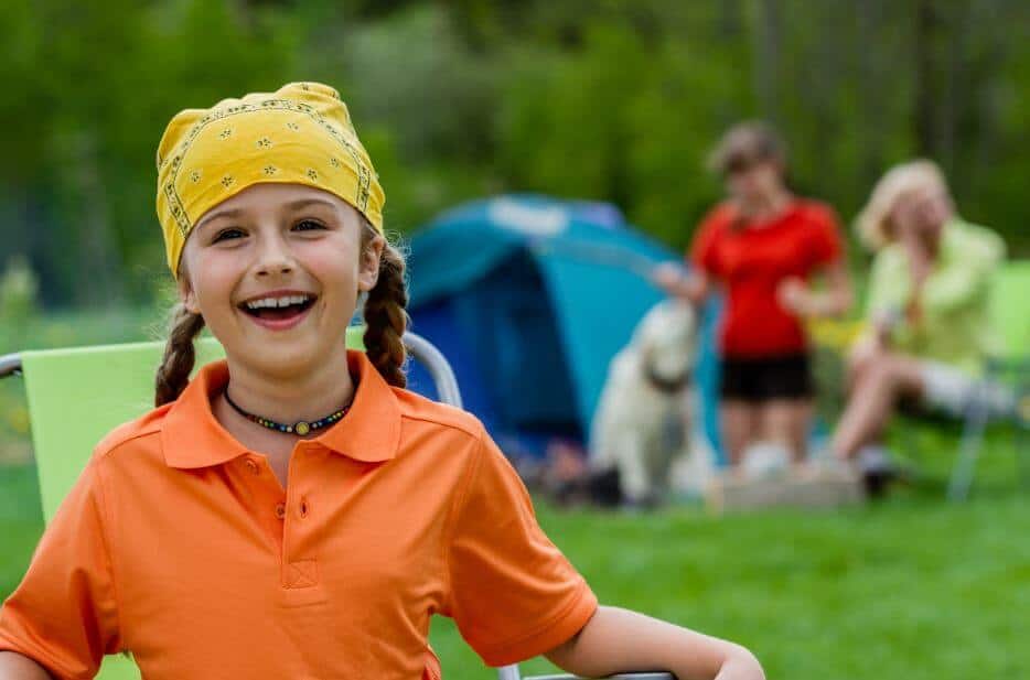 A young girl laughs heartily while camping outside as her family relaxes near the tent in the background.
