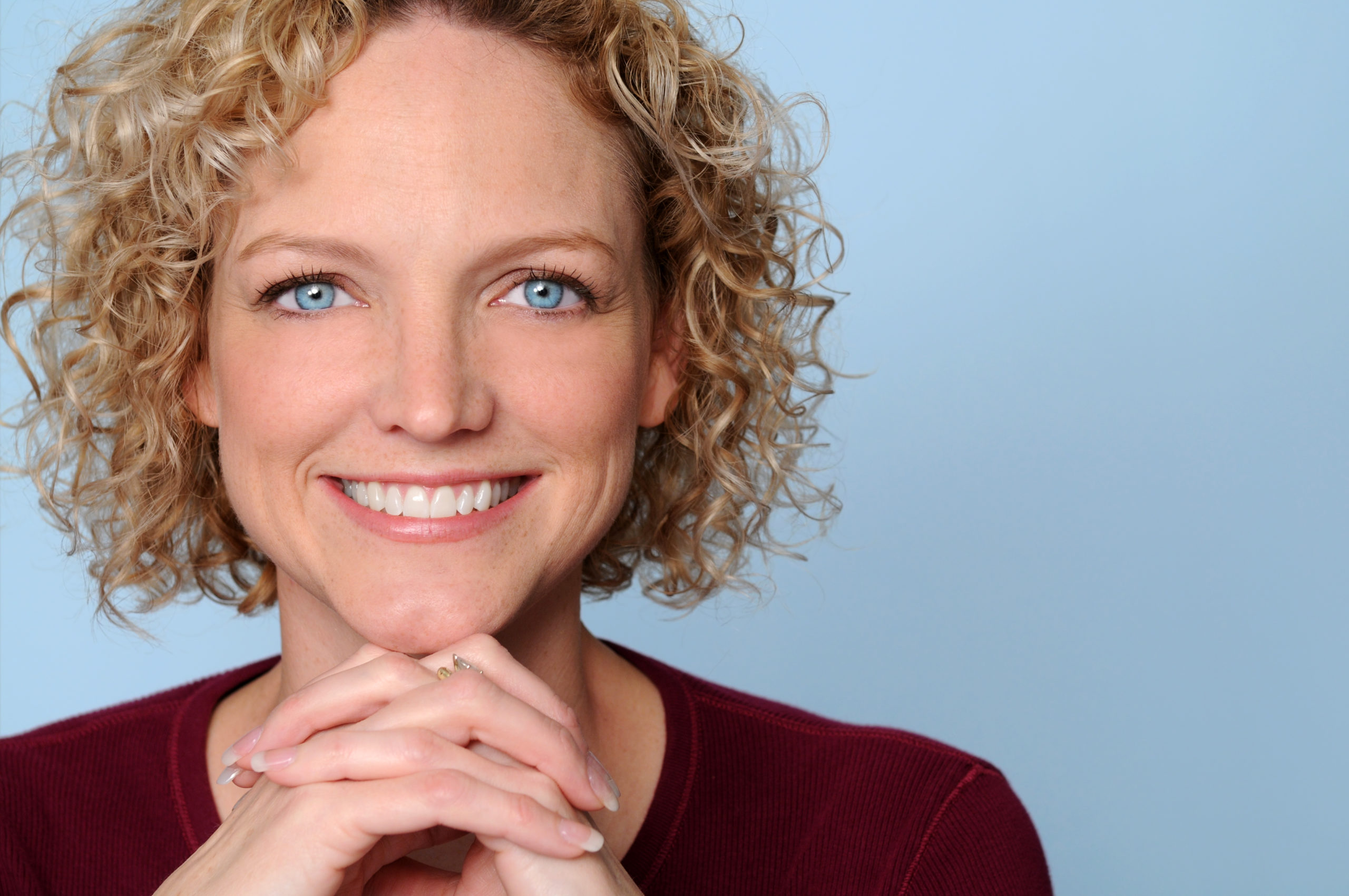 Woman smiling with blonde short curly hair and bright blue eyes, grey background